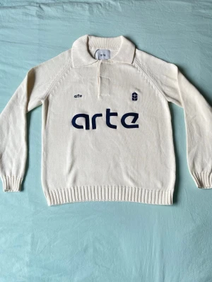 Vit stickad tröja från Arte - Vit stickad tröja från Arte med blå logotyp och text framtill. Tröjan har lång ärm, ribbade muddar och krage med kort knappslå. Perfekt för dig som gillar stilren och sportig design.