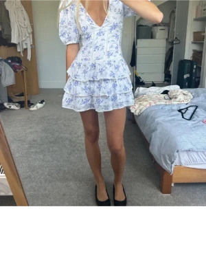 Vit blommig volangklänning - Supersöt kort klänning i vitt med blått blommönster och flera volanglager. Klänningen har v-ringning, puffärmar och markerad midja som ger en feminin siluett. Perfekt för sommarens alla tillfällen!