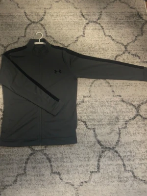 Svart/grå Under Armour zip hoodie - Snygg Under Armour zip hoodie i bästa skick. Clean och en stilren design som passar till allt, perfekt för både vardag och gym. Mjukt och skönt material.