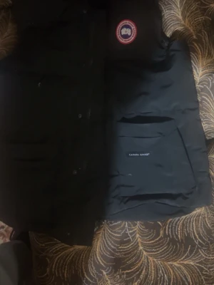 Svart dunväst från Canada Goose - Snygg svart dunväst från Canada Goose med klassisk logga på bröstet och praktiska fickor. Västen är vadderad med dun och har en stilren design som passar perfekt till lager-på-lager. Materialet är slitstarkt och håller värmen under kyliga dagar.