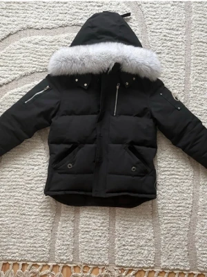 Svart dunjacka från Moose Knuckles - Snygg svart dunjacka från Moose Knuckles med stor huva och lyxig grå fuskpäls. Jackan har flera fickor med dragkedja och tryckknappar samt den ikoniska loggan i metall på ärmen. Perfekt för kalla vinterdagar.
