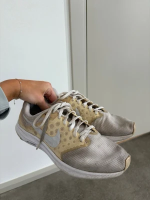 Beige och vita Nike sneakers - Snygga sneakers från Nike i beige och vitt med meshmaterial och vita skosnören. Skorna har en vit sula och den klassiska Nike-loggan på sidan. Perfekta för dig som gillar sportig och avslappnad stil.