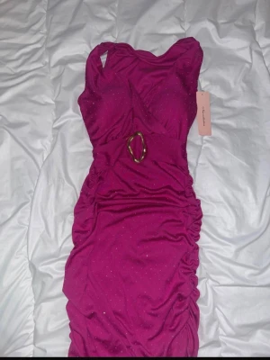 Glittrig rosa omlottklänning - Snygg rosa kortklänning med glittriga detaljer och omlott-design. Klänningen har en markerad midja med ett dekorativt bälte i guld och rynkade sidor för en trendig look. Ärmlös modell som passar perfekt till speciella tillfällen.