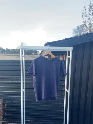 Mörkblå Polo Ralph Lauren t-shirt - Snygg mörkblå t-shirt från Polo Ralph Lauren med klassisk grön logga på bröstet och stor POLO-text i grönt på ryggen. T-shirten har rund hals och korta ärmar, perfekt för en clean och stilren look.