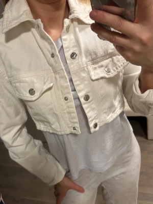Vit cropped jeansjacka från Zara - Snygg vit cropped jeansjacka från Zara med silverfärgade knappar och två bröstfickor med lock. Klassisk krage och raka ärmar. Perfekt för dig som vill ha en trendig och stilren look.