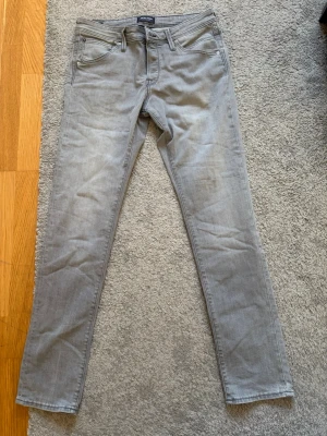 Gråa slim fit jeans från Jack and Jones  - Snygga gråa slim fit jeans från Jack and Jones  med klassisk femficksmodell och diskret vågstickning på bakfickan. Jeansen har normal midja och är tillverkade i mjuk bomullsdenim med lite stretch för extra komfort.