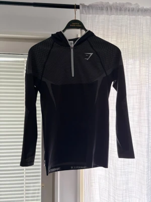 Svart långärmad träningströja Gymshark - Snygg svart långärmad träningströja från Gymshark med halv dragkedja framtill och diskret logga på bröstet. Tröjan har meshdetaljer upptill för extra ventilation och texten 'Be A Visionary' vid nederkanten. Perfekt för gym eller löpning.