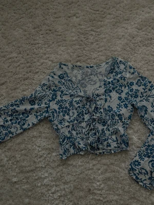 Blommig blus med knytning framtill - Söt blus i vitt med blått blommönster och knytning framtill. Blusen har lång ärm och smockad midja som ger en feminin siluett. Tunt, luftigt material som känns skönt mot huden. Perfekt för en trendig och avslappnad look.