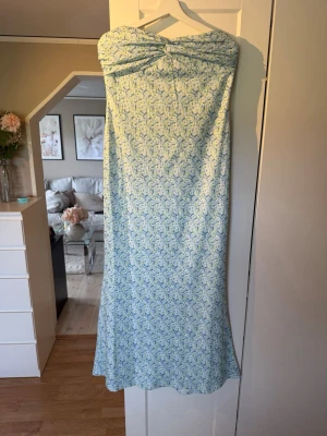 Axelbandslös långklänning - Säljer en ljusblå/ljusgrön axelbandslös långklänning med blommigt mönster och rosettdetalj framtill. Klänningen har en mjuk och flowy passform, lite tyngre material så ingen lätt som blåser upp o syns igenom. Aldrig använd bara testad  