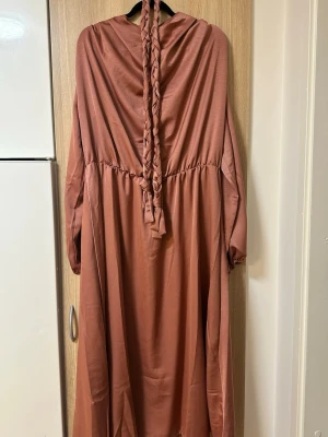 Lång smutsrosa klänning med flätad detalj - Säljer en långklänning i en varm brun ton med långärmad design och en unik flätad detalj baktill. Klänningen har lös passform och är tillverkad i ett mjukt, lätt material som ger ett snyggt fall. Perfekt för dig som gillar stilrena och bekväma plagg.
