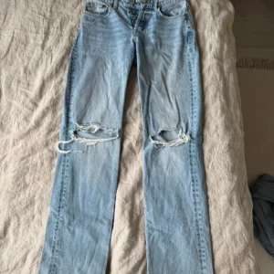 Ljusblå slitna raka jeans - Säljer ett par ljusblå jeans med raka ben och stora slitningar över båda knäna. Jeansen har klassisk femficksmodell och hög midja. Perfekta för en avslappnad och trendig look.