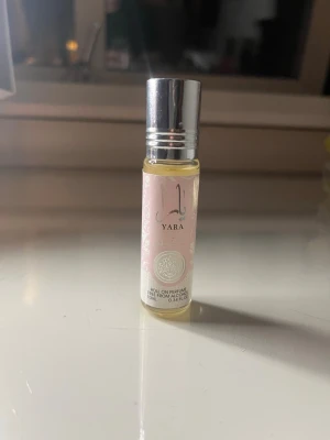 Yara roll-on parfym olja - Säljer en Yara roll-on parfym olja i en liten glasflaska med silverfärgat lock. Flaskan har en ljusrosa etikett med dekorativa mönster och tydlig logga. Perfekt att ha med i väskan för snabb uppfräschning. Aldrig använt 