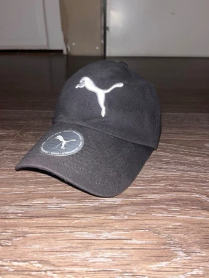 Svart keps från Puma - Snygg svart keps från Puma med den klassiska vita loggan broderad framtill. Kepsen har böjd skärm och justerbar rem baktill för perfekt passform. Tillverkad i bomull för en bekväm känsla och stilren look.