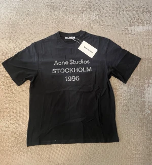 Svart Acne Studios t-shirt Stockholm - Snygg svart t-shirt från Acne Studios med trycket 'Acne Studios STOCKHOLM 1996' på bröstet. Klassisk rund hals och korta ärmar. Tillverkad i mjuk bomull för en skön känsla. Perfekt för dig som gillar minimalistisk och stilren design.
