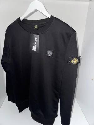 Svart sweatshirt från Stone Island - Svart sweatshirt från Stone Island med klassisk rund hals och ikoniska logopatchar på bröstet och vänstra ärmen. Tröjan har ribbade muddar vid ärmslut och nederkant. Perfekt för en stilren och avslappnad look. Passar jättebra för de som har stolek S.
