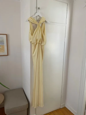 Ljusgul balklänning  - Ljusgul balklänning i ett glansigt material med omlottdesign och knyt vid axlarna. Klänningen har djup v-ringning och ett mjukt, flytande fall som ger en lyxig känsla. 