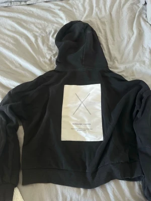 Svart hoodie med tryck på ryggen - Säljer en svart hoodie från The Runaway Collection med stor vit patch och tryck på ryggen. Hoodien har huva med dragsko och breda muddar vid ärmslut och nederkant. Materialet är mjukt och bomullsliknande, perfekt för en avslappnad streetwear-look.