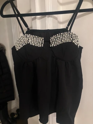 Svart linnetopp med pärldetaljer - Snygg svart linnetopp med tunna axelband och dekorativa pärlor över bysten. Toppen har en figurnära passform upptill och faller löst nedtill. Perfekt för dig som vill sticka ut med coola detaljer.