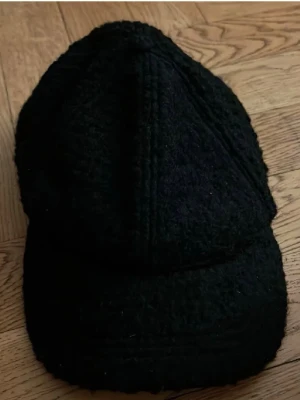 Svart stickad keps - Säljer en svart keps med stickad struktur och böjd skärm. Kepsen har en unik design med paneler och är tillverkad i ett mjukt, värmande material som passar perfekt för kyligare dagar. Inga synliga loggor eller märken.