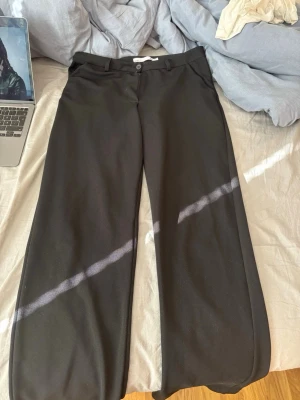 Svarta vida kostymbyxor - Säljer ett par svarta kostymbyxor med vida ben och medel midja. Byxorna har bälteshällor, knapp- och dragkedjestängning samt pressveck framtill. Perfekta för en clean och stilren look.