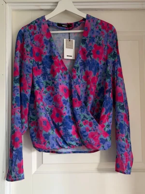 Blommig omlottblus från Bik Bok - Säljer en färgstark omlottblus från Bik Bok i återvunnen polyester. Blusen har ett blommigt mönster i lila, rosa och blå toner, V-ringning och långa ärmar. Perfekt för dig som vill sticka ut med en trendig och bekväm look.