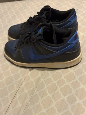 Nike Dunk Low blå och svarta sneakers - Säljer ett par Nike Dunk Low sneakers i blått och svart med vit sula. Skorna har klassisk låg siluett, snörning och den ikoniska swoosh-loggan på sidan. Ovandelen är i läder och textil, vilket ger både stil och komfort. Perfekta för dig som gillar streetwear och sportig look.