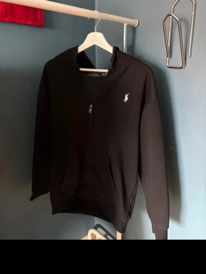 Svart hoodie från Polo Ralph Lauren - Klassisk svart hoodie med dragkedja från Polo Ralph Lauren. Har vit broderad logga på bröstet, huva med snören och känguruficka framtill. Tillverkad i mjukt bomullsmaterial som är skönt att ha på sig. Perfekt för en avslappnad och stilren look.