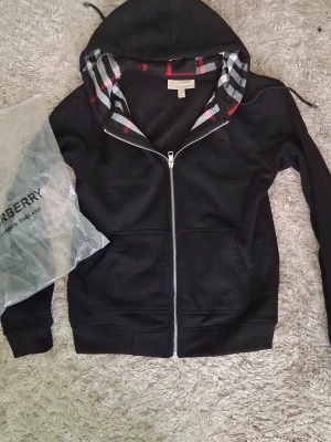 Svart zip hoodie från Burberry - Snygg svart hoodie från Burberry med dragkedja och klassiskt rutigt foder i huvan. Hoodien har fickor framtill och diskret Burberry-logga på bröstet. Perfekt för dig som gillar stilrena plagg med exklusiva detaljer.