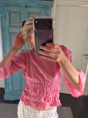 Rosa blus med volang och puffärm - Söt rosa blus med volangdetaljer och puffiga trekvartsärmar. Smockad midja ger en feminin siluett och toppen har en lätt v-ringad rygg. Perfekt för dig som gillar färg och romantiska detaljer. Perfekt nu till sommaren❤️