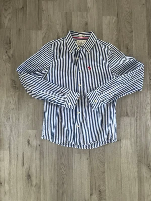 Blåvit randig abercrombie&fitch - Säljer en klassisk blåvit randig skjorta från abercrombie&fitch med lång ärm och knappar framtill. Skjortan har en liten röd logga broderad på bröstfickan och är tillverkad i bomull. Perfekt för en stilren och fräsch look.