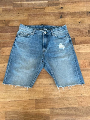 Blå jeansshorts med rå kant - Säljer ett par klassiska blå jeansshorts med rå, fransig kant och slitning på ena låret. Shortsen har fem fickor och knappgylf. Perfekta för en avslappnad stil i sommar.