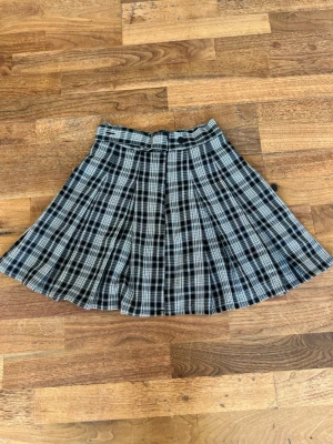 Svartvit rutig kortkjol med bälte - Snygg svartvit rutig kortkjol med plisserad design och ett dekorativt bälte i midjan. Kjolen har knäppning framtill och passar perfekt till en trendig vardagslook. Klassisk och tidlös stil som är lätt att matcha.