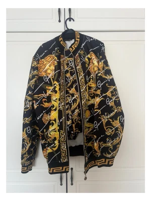 Versace jacka och byxor  - Snygg svart bomberjacka från Versace med ikoniskt barockmönster i guld och vitt. Jackan har ribbade muddar, dragkedja framtill och detaljer med grekiskt mönster. Fodrad insida och signaturtryck över hela plagget. Perfekt för dig som vill sticka ut.