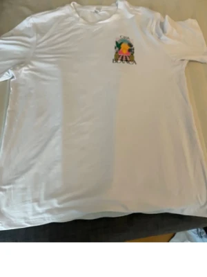 Vit Casa Blanca t-shirt med tryck - Vit t-shirt från Casa Blanca med färgglatt grafiskt tryck på ryggen som visar en solnedgång, palmer och texten 'Casa Blanca'. Klassisk passform och rund halsringning. Perfekt för dig som gillar stilrena plagg med en twist.