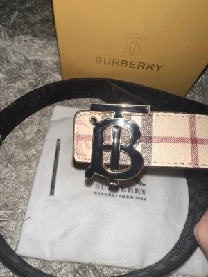 Burberry bälte - Snyggt Burberry bälte med det ikoniska beigea och röda rutmönstret samt en stor metallspänne med Burberrys logotyp. Bältet har detaljer i svart och beige och är tillverkat i skinn. Perfekt accessoar för att lyfta din outfit.