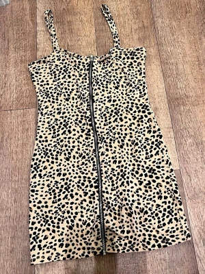 Leopardmönstrad kort klänning - Säljer en snygg leopardmönstrad kort klänning med smala axelband och dragkedja framtill. Klänningen har en beige bas med svarta och bruna fläckar, figurnära passform och ärmlös design. Perfekt för dig som vill sticka ut med ett djurmönster!