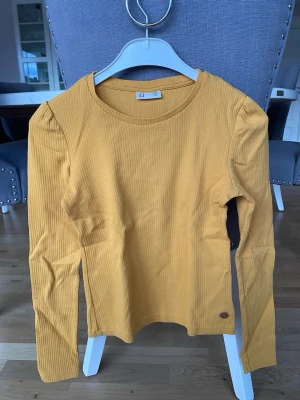 Gul ribbad långärmad topp från Ü - Snygg gul ribbad topp från Ü med lång ärm och rund halsringning. Toppen har en liten logodetalj nertill och är tillverkad i mjuk bomull. Perfekt för dig som gillar en enkel men trendig stil. Köparen står för frakt.