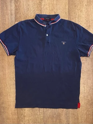 Mörkblå Piké från Gant - Klassisk marinblå pikétröja från Gant med korta ärmar och vit-röd-blå randiga detaljer på krage och ärmslut. Liten broderad logga på bröstet. Tillverkad i mjuk bomull för skön känsla. Perfekt till jeans eller shorts. Storlek Medium. Snabb Leverans!