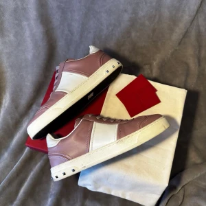 Valentino Garavani flycrews rosa/vit - Snygga sneakers från Valentino Garavani i en kombination av ljusrosa och vitt läder. Skorna har klassisk låg profil, vita partier på sidorna och coola nitar längs sulkanten baktill. Allt originellt tillkommer. Köpta för 5500kr. Storlek 37 men passar 38. En extra ni tillkommer samt. En bit har tappats.