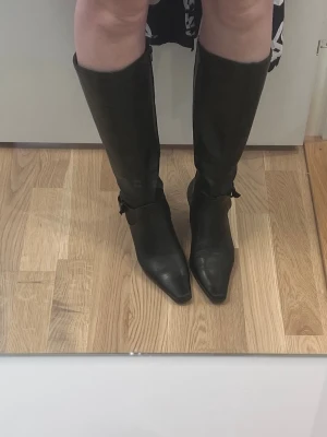 Svarta knähöga boots med klack - Säljer ett par svarta knähöga boots i skinnimitation med spetsig tå och smal låg klack. Stövlarna har ett dekorativt spänne vid ankeln och en elegant, stilren design som passar perfekt till kjol eller klänning.