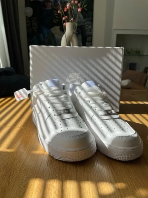 Nike Air Force 1 vita sneakers - Klassiska vita Nike Air Force 1 sneakers med låg profil och perforerad tåbox för extra ventilation. Skorna har en chunky sula, vita skosnören och diskret Nike-logga på sidan. Tillverkade i slätt läder med detaljer i textil på plösen.