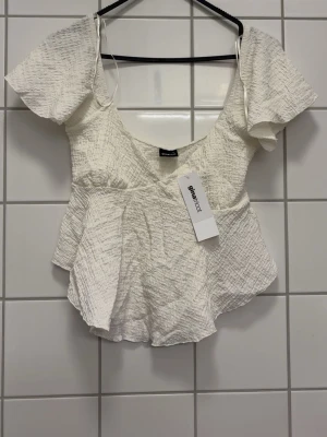 Vit blus med volang - Supersöt vit blus med v-ringning, korta vida ärmar och volang nedtill. Tillverkad i ett lätt, strukturerat tyg som ger en luftig känsla. Perfekt för dig som vill ha en feminin och trendig topp till sommaren.