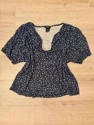 Blommig blus med drapering - Söt blommig blus från Lindex i svart med små blå blommor. Blusen har korta, lite puffiga ärmar och en djupare draperad ringning. Materialet känns lätt och luftigt, perfekt för varmare dagar. Passformen är normal och toppen ger en feminin siluett.