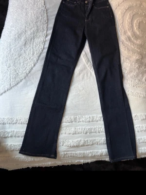 Mörkblå raka jeans - Säljer ett par mörkblå jeans med rak passform och hög midja. Klassisk femficksmodell med knapp och dragkedja framtill. Jeansen har en stilren look och är tillverkade i denim, perfekt för en clean och enkel outfit.