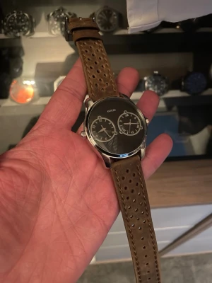Guess Dual Time klocka  - Stilren klocka från Guess med svart urtavla och två separata tidvisare. Klockan har ett perforerat brunt läderband och boett i rostfritt stål . Perfekt accessoar för dig som gillar en sportig men ändå klassisk look.
