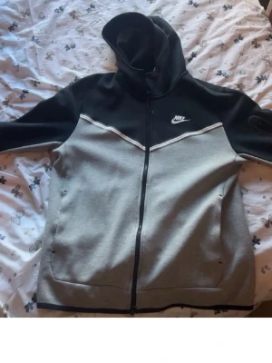 Nike tvåfärgad hoodie med dragkedja - Snygg hoodie från Nike i svart och grått med vit logga på bröstet. Tröjan har hel dragkedja framtill, två fickor och en stor huva. Tillverkad i mjukt sweatshirtmaterial, perfekt för en avslappnad streetwear-look.