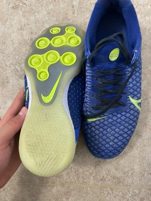 Blå Nike fotbollsskor med neon detaljer - Säljer ett par blå Nike fotbollsskor med cool struktur på ovandelen och svarta skosnören. Sulan har unika neon-gula cirkulära mönster och Nike-logga. Skorna är designade för bra grepp och kontroll på planen. Perfekta för dig som vill sticka ut på matchen.