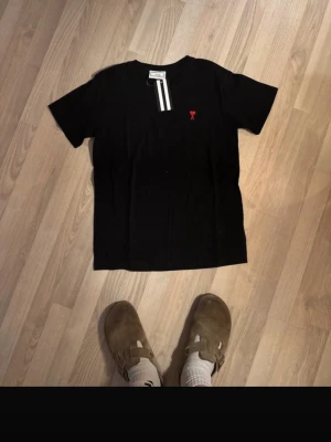 Svart t-shirt från Ami Paris - Svart t-shirt från Ami Paris med korta ärmar och en liten röd logga broderad på bröstet. Tröjan är köpt i Grekland, och tröjan är flitigt använd (8/10 skick) därav det billiga priset. Obs tagsen är borttappade. 100% cotton.
