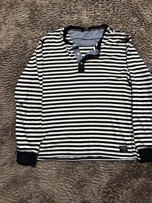Randig långärmad tröja Lindbergh Blue - Snygg långärmad tröja från Lindbergh Blue med mörkblå och vita horisontella ränder. Tröjan har knappar vid halsen och mörka muddar vid ärmsluten. Perfekt för en avslappnad och stilren look.