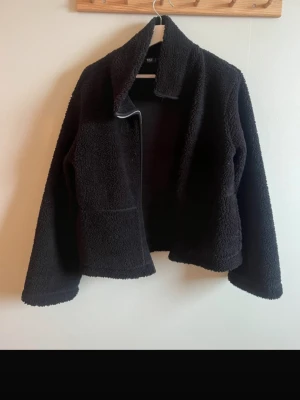 Svart teddyjacka från 157 - Mysig svart teddyjacka från 157 med bred krage och mjukt, fluffigt material. Jackan har en rak passform och är perfekt för kyliga dagar. Enkel design utan synliga knappar eller dragkedja framtill.